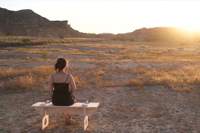 A visitor sits on Rose Hallgren's wooden bench overlooking the desert hills at sunset, seen from behind.