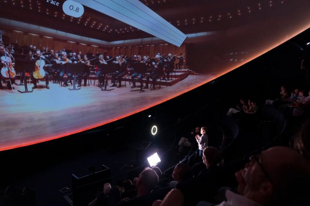 A child stands at the front of the dome beneath a projected orchestra and tempo indicator, with the audience seated nearby.