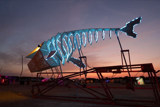 A low side view of the whale with cyan ribs and a raised tail against the evening sky, its metal mechanism visible underneath.