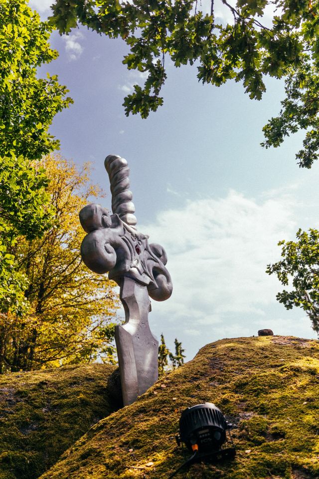 A pale sculpture of coiled forms stands on a mossy granite outcrop under an oak, with a stage light at its foot.
