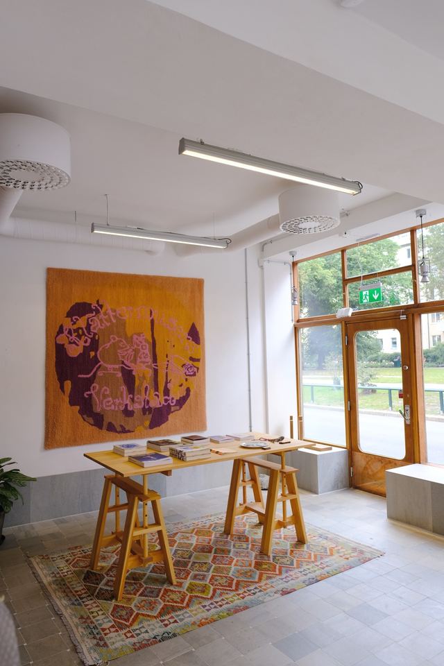 The finished rug hangs above a table of books in Klättermusen's Verkstad, with the park visible through the open door.
