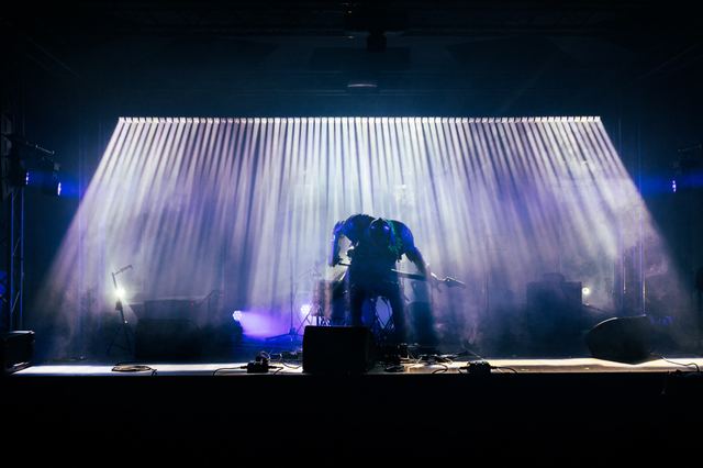 Beams of light rake through haze across a draped backdrop, a figure silhouetted at the microphone in front of it.