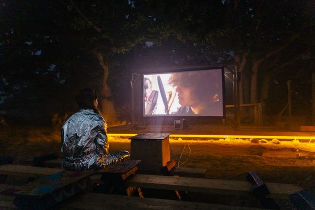 One person sits alone on a bench in front of the outdoor cinema screen at night, the ground lit yellow in front of it.