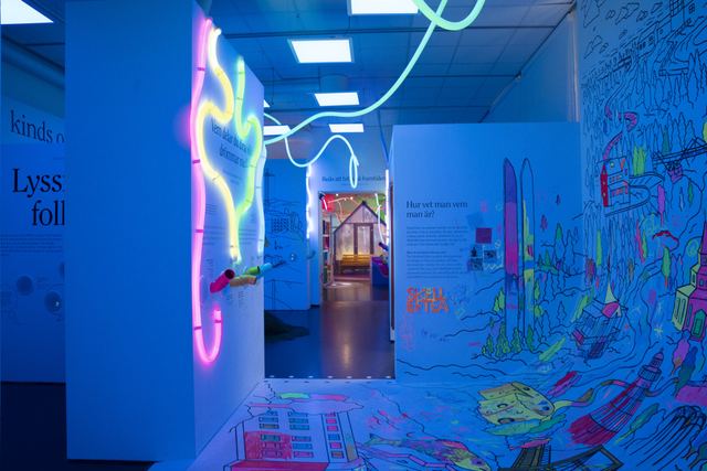 Blue light fills a passage between a listening wall and a hand-coloured mural, with luminous lines continuing across the ceiling.