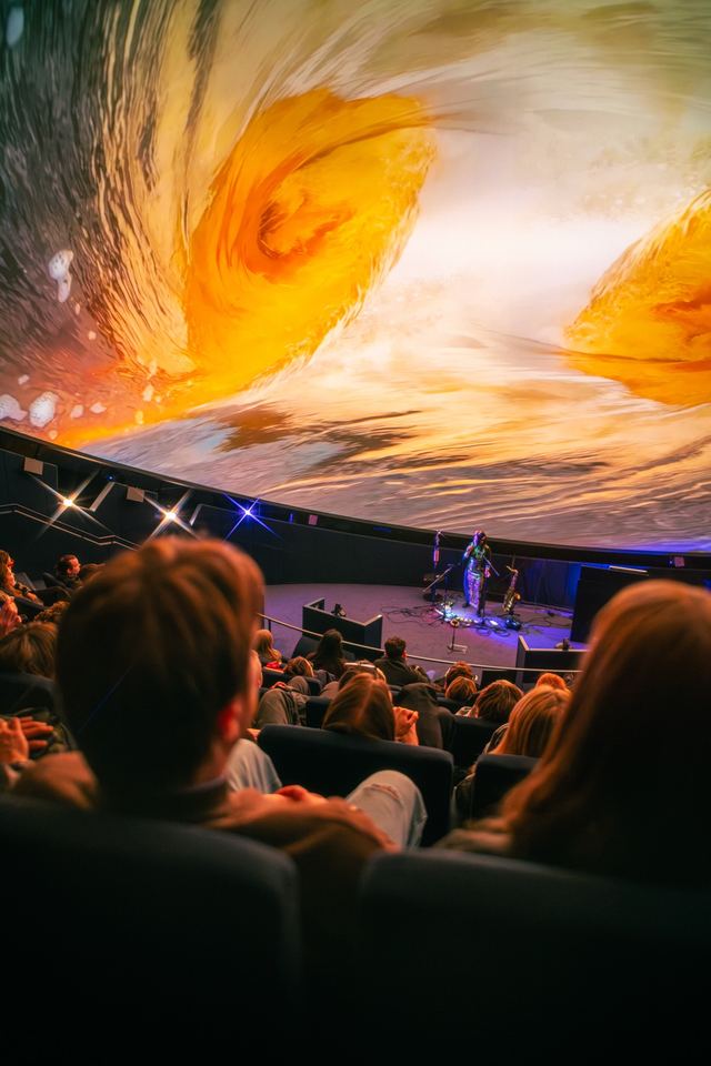 An audience reclines beneath a dome filled with swirling gold and white imagery during a live performance.