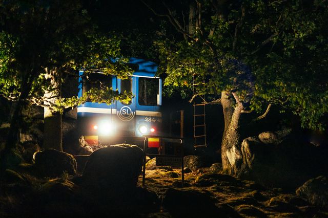 A truck parked among trees at night, its headlights and a blue-lit cabin glowing through the branches.
