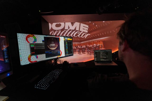 A developer works at a laptop and a monitor showing the dome projection setup, with the orchestra projected in the background.