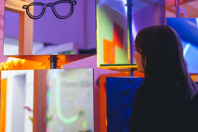 A visitor looks into reflective display boxes edged with orange and pink light.