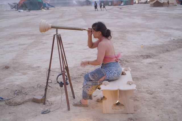 A visitor sits on a small wooden bench and looks through WYSIWYG in an open desert setting.