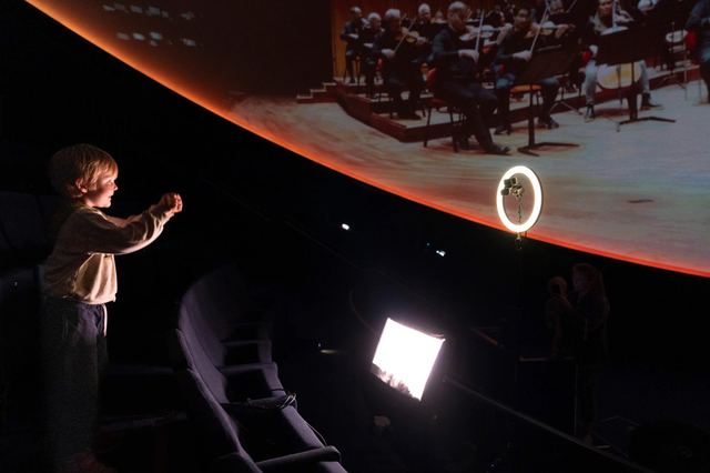 A child raises both arms beneath the projected orchestra, facing a tracking camera mounted inside a ring light.