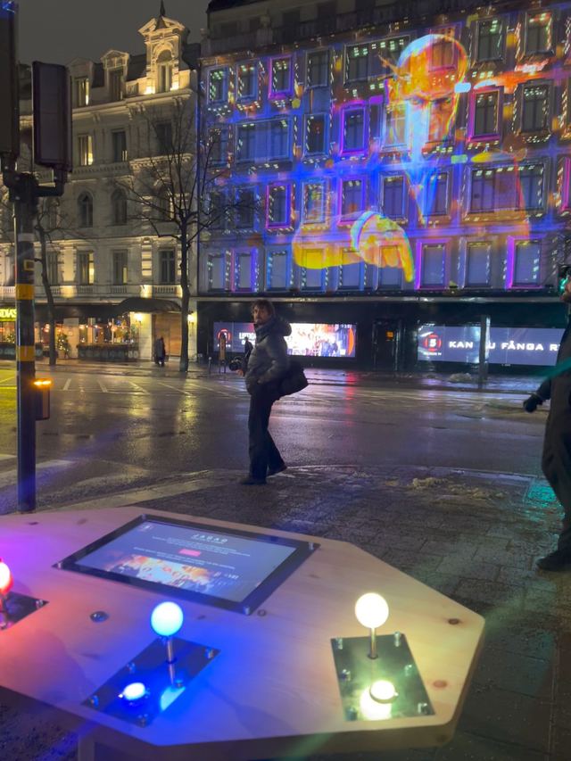 Glowing arcade joysticks face a Stockholm building covered in colourful game projections across the wet street.