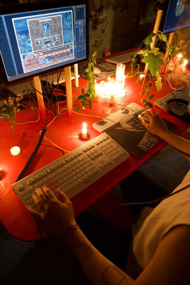 An overhead view of a player's hands at the keyboard and mouse, with Heroes of Might and Magic III on the screen and candles between the stations.