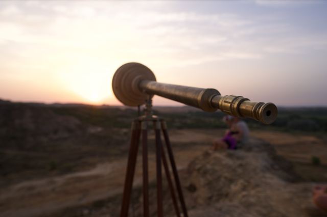 A brass telescope with a circular wooden housing stands on a tripod against a warm desert sunset.