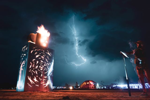 A giant patterned lighter burns in an open field at night as a lightning bolt illuminates the sky behind it.