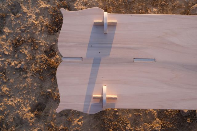 A view down onto the bench's plywood seat, showing its curved cutouts, wooden pegs and long evening shadows.