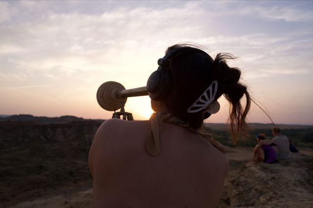 A visitor wearing headphones looks through the telescope as the sun sets over the landscape.
