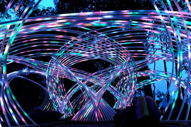 Looking up from inside Kagora, multicoloured light curves cross above visitors beneath the evening sky.