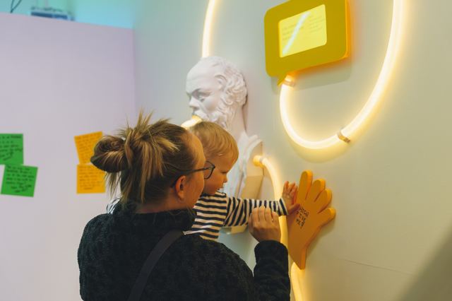 An adult holds a child reaching for a yellow hand-shaped touch panel, beside a glowing loop of light and a white bust.