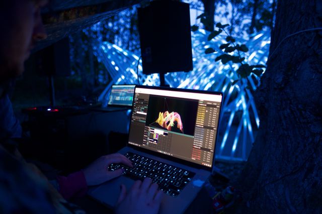 Hands at a laptop showing a digital model of Kagora, with the illuminated pavilion glowing blue among the trees behind it.