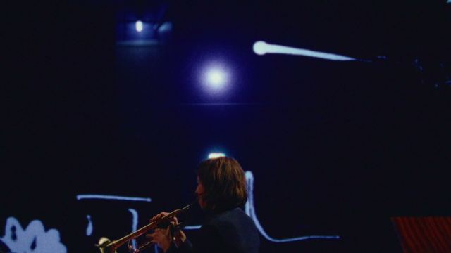 Adam Sass plays trumpet in blue light, a white line of the score curling across the wall behind him.
