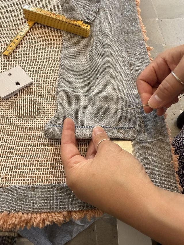 Hands stitch the linen backing along the rug's edge beside a folding ruler and mounting clip.