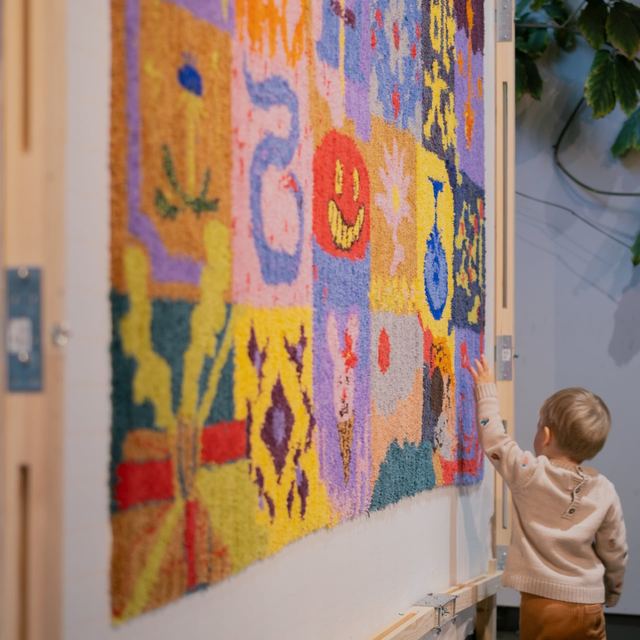 A child reaches towards a large patchwork of colourful tufted textiles, including flowers, figures and a smiling face.