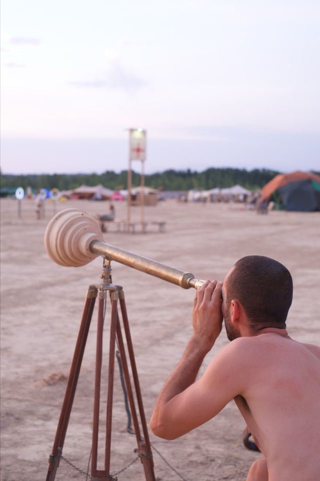 A visitor looks through the brass telescope at dusk, seen from behind with its wooden housing and tripod in view.