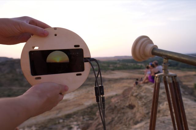 The opened circular wooden housing reveals the phone and its round landscape view beside the telescope.