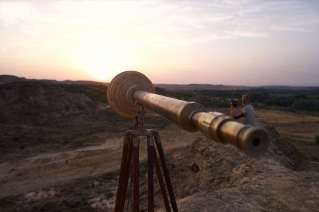 A close view along the brass telescope towards its circular wooden housing and the setting sun, with two visitors sitting in the distance.