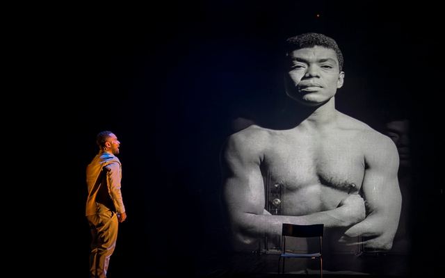A performer faces a towering black-and-white portrait projected onto the dark stage.