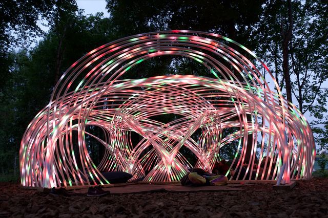 A frontal view of Kagora at dusk, with pink, green and white light tracing the woven arches above visitors resting on cushions.