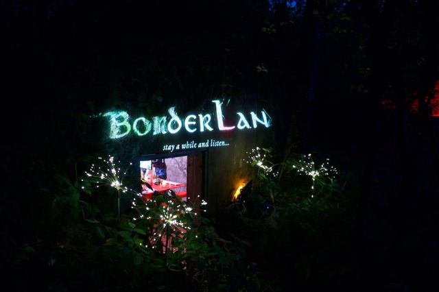 An illuminated BorderLAN sign above the cellar entrance reads 'stay a while and listen', surrounded by foliage and small lights.