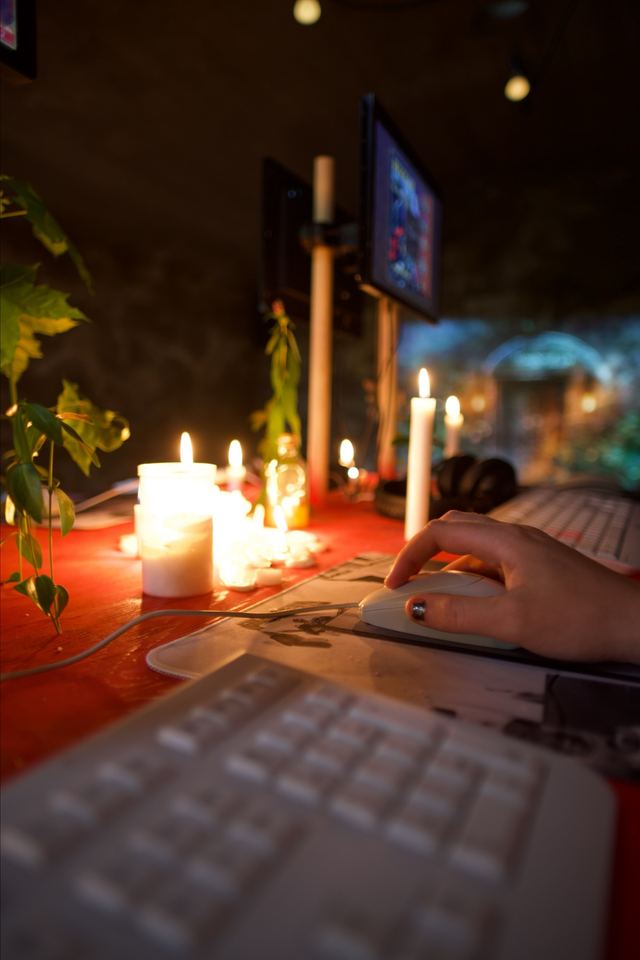 A hand rests on a white mouse beside candles and plants, with a game screen and the projected cellar doorway in the background.