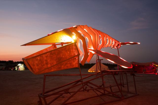 The whale's open mouth, ribs and near fin glow orange and red against a pink evening sky.